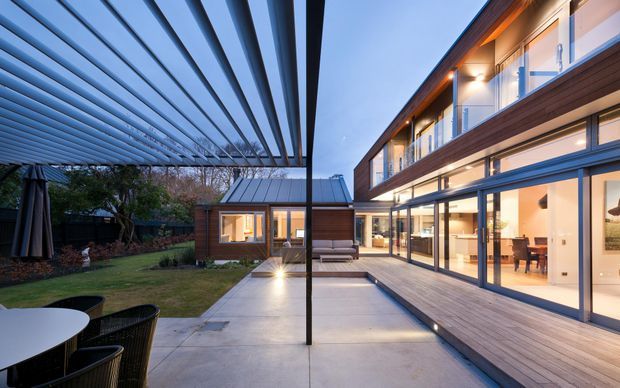 Contemporary two-storey residence viewed from a landscaped courtyard terrace at dusk. The home combines dark timber cladding, extensive floor-to-ceiling glazing, upper-level glass-balustraded balconies, and a broad outdoor pergola with adjustable horizontal louvres. A paved patio, timber decking, outdoor seating, dining furniture, and warm interior lighting create a connection between the courtyard and the open-plan interior.