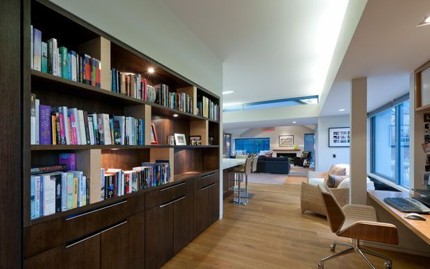 Open-plan contemporary interior featuring a large dark timber built-in bookcase with integrated display lighting and lower cabinetry in the foreground. A timber-floored living area, dining or kitchen zone, and compact home office with desk and chair extend beyond, with extensive glazing and a skylight bringing in natural light.