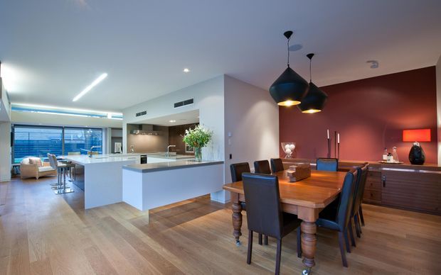 Open-plan contemporary kitchen, dining and living area with a large white island, integrated cabinetry, timber flooring, a substantial timber dining table with dark upholstered chairs, and a warm feature wall with pendant lighting and sideboard.