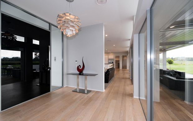 A bright contemporary residential entry hall with pale timber flooring, white walls, extensive floor-to-ceiling glazing, and a black-framed glass door. A sculptural pendant light hangs above a slim console table, while the hallway leads toward an open-plan kitchen and living area. Outdoor greenery and seating are visible through the glazing and reflections.