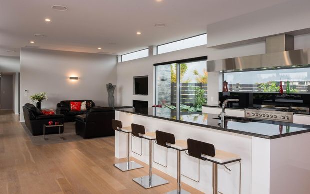 Open-plan contemporary kitchen and living area with a white island incorporating a raised breakfast bar and four dark upholstered stools. The kitchen features dark stone countertops, integrated stainless-steel appliances, extensive glazing, and sleek white cabinetry. A black sectional sofa, media unit, timber flooring, recessed lighting, and neutral walls define the adjoining living room.