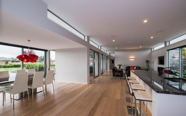A bright, open-plan contemporary interior combining a dining area, kitchen, and living zone. Full-height glazing and clerestory windows bring in abundant daylight, while timber flooring runs throughout. The kitchen features a long island with a dark stone benchtop, white cabinetry, and bar seating. A vivid red pendant light provides a strong focal accent above the dining table.