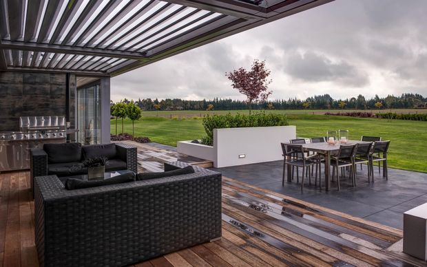 A covered outdoor entertaining area with a contemporary pergola featuring adjustable louvred slats, dark woven outdoor lounge furniture, and a dining table on a large paved terrace. Timber decking transitions to grey paving, with a raised white rendered planter wall and expansive landscaped lawn and rural fields beyond.