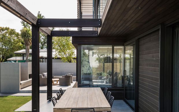 Contemporary outdoor dining terrace beside a dark-clad modern home, featuring a long timber dining table, black metal chairs, exposed steel pergola framing, extensive glazing, and a landscaped courtyard with a secondary seating area.