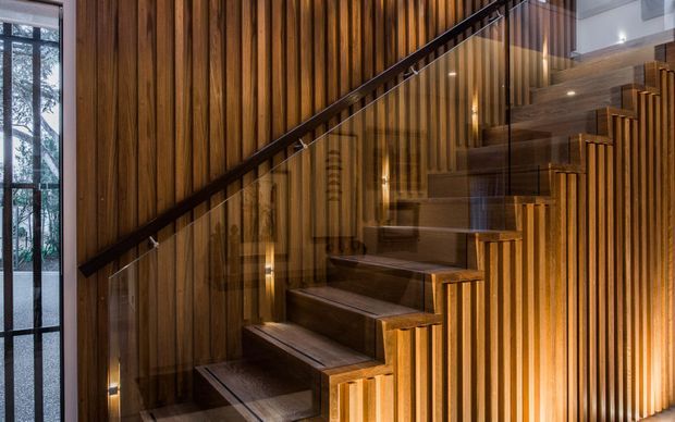 A dramatic timber-lined stairway with dark wood treads and a slim dark handrail, enclosed by vertical timber battens. Warm integrated lighting highlights the stair and wall surfaces, while glass balustrading and a tall glazed opening bring in natural light and views of trees.