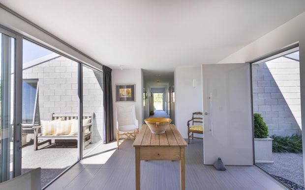 Bright, minimalist open-plan interior with a timber dining table and chairs, white walls, pale flooring, and extensive floor-to-ceiling glazing. The glazing opens onto a small paved outdoor courtyard or patio, while a hallway leads deeper into the home. Natural light, restrained furnishings, and neutral finishes create an airy, calm atmosphere.