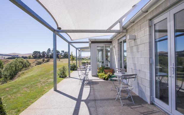 A long, covered outdoor terrace running alongside a contemporary building, with a concrete-paver floor, grey masonry wall, glazed French doors and windows, and a lightweight steel-framed canopy with translucent roofing. Small café tables and chairs sit along the terrace, overlooking an open rural landscape.