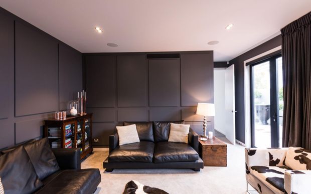 A compact, moody media or living room with dark charcoal wall panelling, a black leather sofa and armchair, a central low coffee table, a timber side cabinet, and a cowhide-patterned occasional chair. Full-height dark curtains frame glazed double doors, while recessed ceiling lights and a table lamp provide warm illumination. The room has a refined contemporary feel with strong tonal contrast and layered textures.