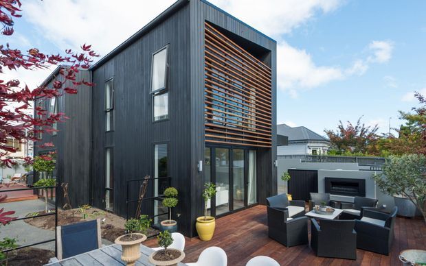 Contemporary multi-level dark-clad residential exterior with a tall glazed opening screened by horizontal timber louvres. A timber deck with outdoor lounge furniture, potted plants, and sculpted landscaping occupies the foreground, while neighbouring suburban homes are visible beyond.