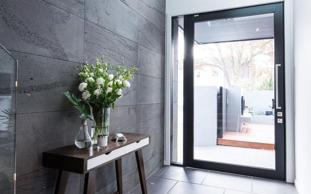 A bright contemporary entryway with a dark grey textured concrete feature wall, large-format grey floor tiles, and a slim timber console table topped with flower arrangements. A black-framed glazed door opens to a small outdoor courtyard or terrace, bringing in strong natural light.