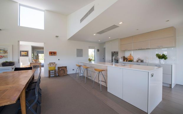 Bright, spacious contemporary open-plan kitchen and dining area with a large white island, integrated appliances, pale timber overhead cabinetry, glossy white splashback, and timber dining table. High ceilings, clerestory-style glazing, recessed lighting, and neutral finishes create an airy, clean interior.