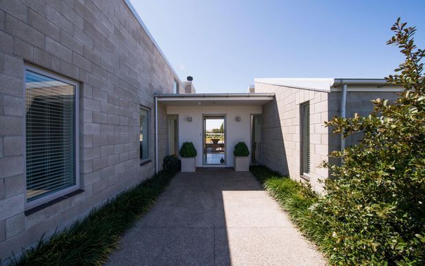 A sheltered entry courtyard between low, rectilinear buildings clad in pale grey concrete masonry blocks. A covered passage leads to a glazed entrance door, flanked by two potted topiary plants, with narrow planted borders and mature shrubs along the textured concrete path. Horizontal window blinds and simple metal downpipes reinforce the clean, functional exterior design.