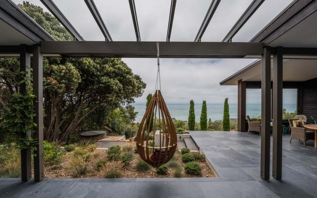 A covered outdoor terrace or courtyard framed by dark timber posts and a translucent roof, with a suspended egg-shaped hanging chair in the foreground. The space opens onto landscaped garden beds and a paved terrace beside a modern house, with coastal water visible in the distance. The palette is dark timber, grey paving, and muted natural planting.