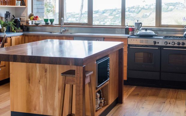 A warm, timber-rich kitchen with a large central island, integrated open shelving and beverage storage, stainless-steel sink and appliances, and a large black range cooker. Timber-framed windows provide expansive views over a hilly residential landscape. The kitchen has a practical, inviting character with natural wood cabinetry and flooring.