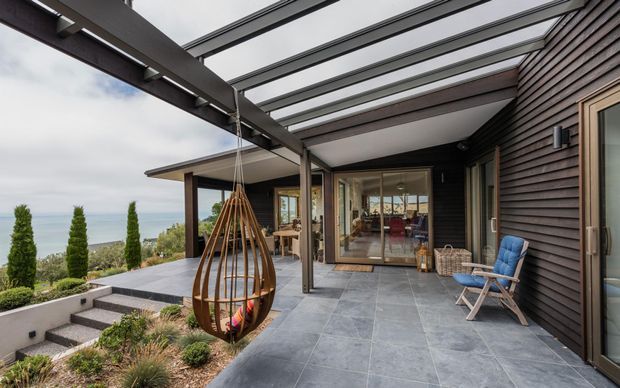 A contemporary covered outdoor terrace overlooking a coastal landscape. The space features large grey stone-look paving, dark horizontal timber cladding, a steel-framed translucent pergola, frameless or expansive glazing, an outdoor hanging pod chair, and a blue lounge chair. Steps lead down through landscaped planting, with the living area visible through the glazed doors.