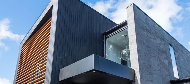 Contemporary multi-level building exterior viewed from below, featuring dark vertical cladding, a projecting balcony with dark metal fascia, a tall glazed section, a prominent horizontal timber louvre screen, and pale large-format stone cladding. The angular composition and crisp rooflines create a bold modern facade against a partly cloudy sky.