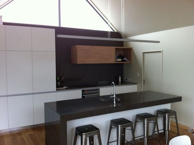 A contemporary kitchen with a large dark stone island and integrated sink, accompanied by four metal stools. The room features tall white flat-panel cabinetry, dark splashback and benchtops, a timber wall-mounted storage unit, timber flooring, and high clerestory glazing beneath a sloped ceiling.