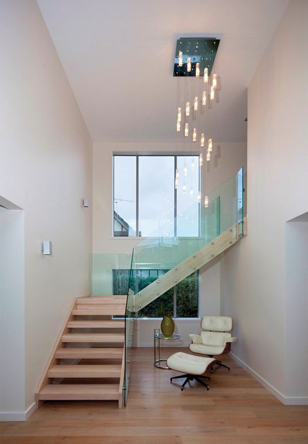 Bright contemporary stair hall with an open timber staircase, clear glass balustrades, white walls, and warm timber flooring. A sculptural multi-drop pendant chandelier hangs from a ceiling recess above the stair, while a cream lounge chair and ottoman sit beneath the landing beside large windows.