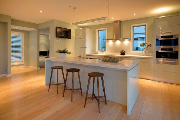 A bright, open-plan contemporary kitchen with a large pale island, integrated white cabinetry, stainless-steel appliances, pendant lighting, and three timber bar stools. Light timber flooring, pale walls, recessed lighting, and windows create a warm, spacious feel. A living area and hallway are visible beyond the kitchen.