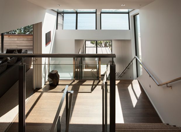 A bright, modern multi-level interior landing or stair hall with extensive floor-to-ceiling glazing, white walls, timber flooring, and dark metal-and-glass balustrades. A stair descends in the foreground toward a central lower-level void, with a living area and television visible to the left. Strong sunlight creates geometric shadows across the floor.