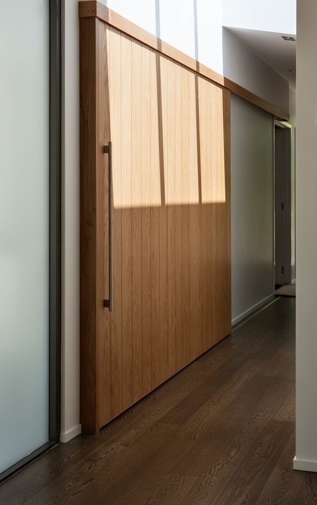 A contemporary interior hallway featuring a large full-height timber pivot door with vertical wood panelling and a long dark metal handle. The door is set within white walls beside tall glazing, with warm dark timber flooring continuing through the corridor. Strong sunlight creates a horizontal band of light and shadow across the door, while recessed ceiling lighting and additional doorways extend into the background.