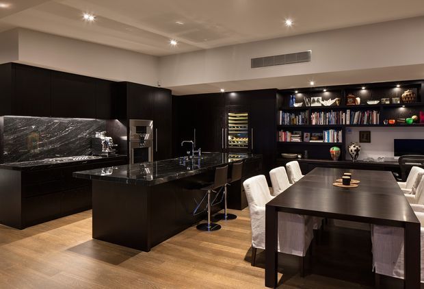 A sophisticated open-plan kitchen and dining area with black built-in cabinetry, a large central island, dark stone benchtops and splashback, integrated appliances, and a built-in display bookshelf with wine storage. A substantial dark dining table with upholstered chairs occupies the foreground. Recessed ceiling lighting creates a warm, refined atmosphere, while timber-toned flooring adds contrast.