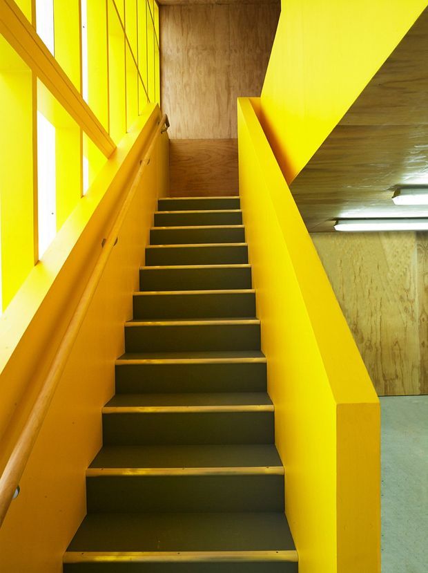 A bold interior stairway with dark timber treads enclosed by intensely yellow-painted timber side walls, balustrades, and window framing. Timber wall and ceiling surfaces continue around the landing, while fluorescent strip lighting is visible in the lower adjoining area. The composition is highly graphic, geometric, and industrial in character.