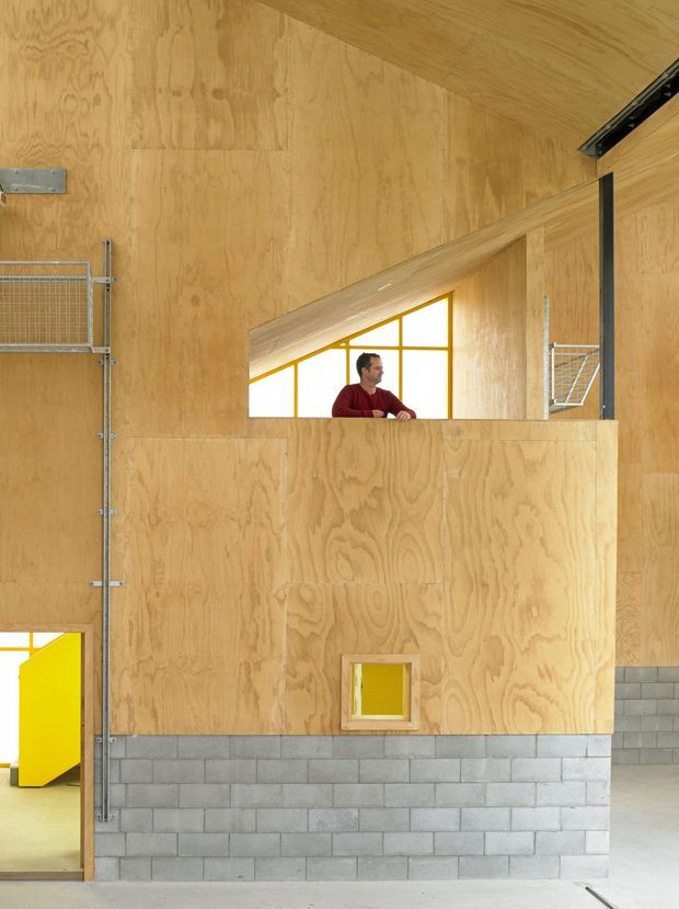 Interior view of a large industrial or workshop-like space with exposed plywood wall and ceiling surfaces, a lower wall of grey concrete masonry blocks, bright yellow framed openings and doors, and utilitarian galvanized metal mesh elements. A small upper-level opening reveals a person and a yellow-framed glazed window. The space is open, functional, and brightly lit with a strong raw-material palette.