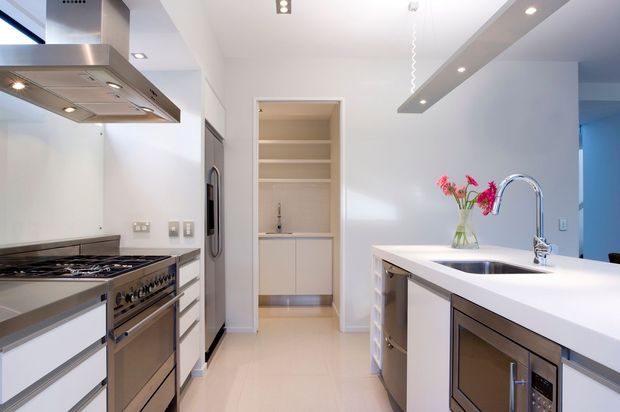 Bright contemporary kitchen with a large white island and integrated sink, stainless-steel range and appliances, extensive white cabinetry, recessed lighting, and a built-in pantry visible at the rear. The space has clean lines, a neutral palette, and a moderately spacious layout.