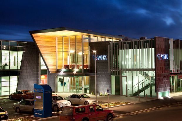 Night-time exterior view of a contemporary multi-storey commercial building, with a dramatic projecting angular canopy clad in warm timber, extensive floor-to-ceiling glazing, dark tiled or panelled facade sections, and illuminated corporate signage. The building fronts a street with parked vehicles and a landscaped entry forecourt.