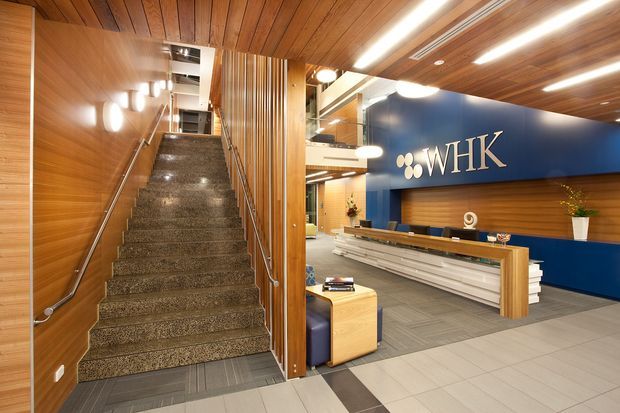 A polished commercial reception and stairway interior. The foreground features a reception desk with a large WHK logo wall behind it, while a carpeted stair rises beside warm timber wall and ceiling linings. Vertical timber battens, glass balustrades, recessed linear lighting, grey carpet tiles, and large light-grey floor tiles create a contemporary corporate fit-out.