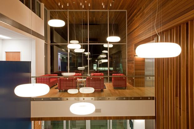 Interior view of a contemporary commercial lounge or reception space, featuring a dramatic double-height glazed wall with mirrored reflections, red upholstered seating, a timber-lined ceiling and feature wall, and numerous suspended oval pendant lights. A raised mezzanine-like platform or balcony edge overlooks the lower level, with visible glazing and a polished timber floor.
