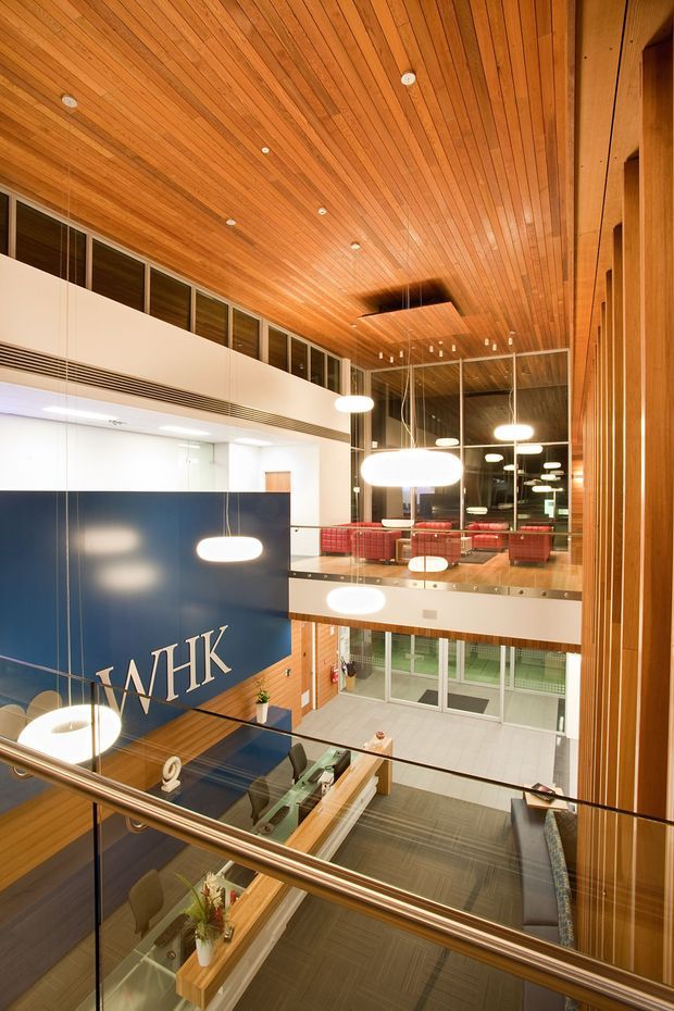 A tall, contemporary commercial interior viewed from an upper-level landing or mezzanine. The space features extensive warm timber panelling across the ceiling and vertical walls, a double-height atrium with glass balustrades, suspended oval pendant lights, dark blue branded wall signage reading “WHK,” reception or collaborative work areas, and a visible upper lounge with red seating. The open void, multiple levels, and extensive glazing create a bright, polished corporate workplace atmosphere.