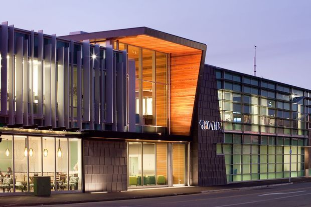Twilight exterior photograph of a contemporary commercial building with a layered facade. The building combines dark textured cladding, extensive gridded glazing, vertical metal fins, and a warm timber-lined recessed upper volume. Ground-floor windows reveal illuminated interior work and dining areas, while a street frontage runs across the foreground.