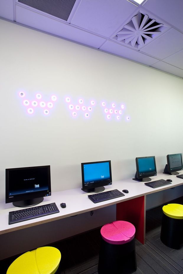 A compact computer workspace with a row of desktop computers, keyboards, and mice arranged along a long white counter. Brightly coloured round stools sit beneath the workstations, while a decorative cluster of glowing circular wall lights adds a playful accent. The room has a suspended acoustic ceiling with visible ventilation grilles and dark striped carpet flooring.