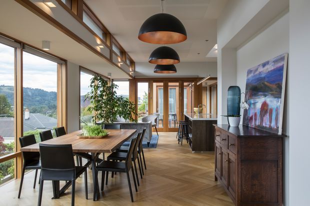 A bright, contemporary open-plan dining and kitchen area with expansive timber-framed glazing overlooking a leafy suburban landscape. The room features a large timber dining table with black chairs, a dark kitchen island, herringbone timber flooring, dark wood sideboard cabinetry, white walls, and a series of oversized black pendant lights with warm copper interiors. A colourful abstract artwork and indoor planting add visual interest.