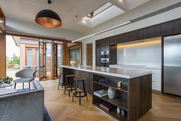 A spacious, refined contemporary kitchen with a large dark timber island, white stone benchtop, integrated stainless-steel appliances and open shelving. Three stools line the island, while a lounge seating area and glazed doors to an outdoor space are visible beyond. A skylight, concealed linear lighting and a sculptural black-and-copper pendant provide strong architectural and decorative features. The timber floor is laid in a herringbone pattern.