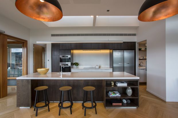 A spacious contemporary kitchen with a large island featuring a pale stone benchtop, dark timber cabinetry, integrated appliances, open shelving, and three bar stools. The room has herringbone timber flooring, white walls, skylights, linear air conditioning vents, and striking oversized black-and-copper pendant lights. Glazing and a timber-framed doorway open toward an exterior balcony or terrace.