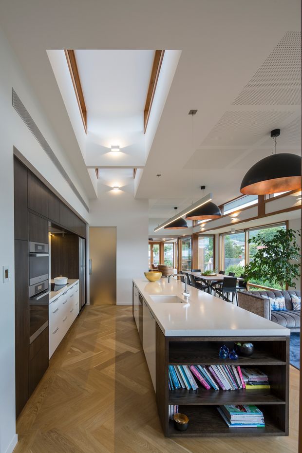 A bright, open-plan contemporary kitchen and living area viewed along a large central island. The kitchen features dark timber-toned full-height cabinetry, integrated appliances, white drawers, and a broad white benchtop with an integrated sink. The island end has dark open shelving filled with books and decorative objects. Herringbone timber flooring runs through the space, while large windows and glazed doors bring in natural light. Black dome pendant lights, timber-framed glazing, a long skylight, and perforated acoustic ceiling panels add architectural detail. A dining and lounge area is visible beyond the island.