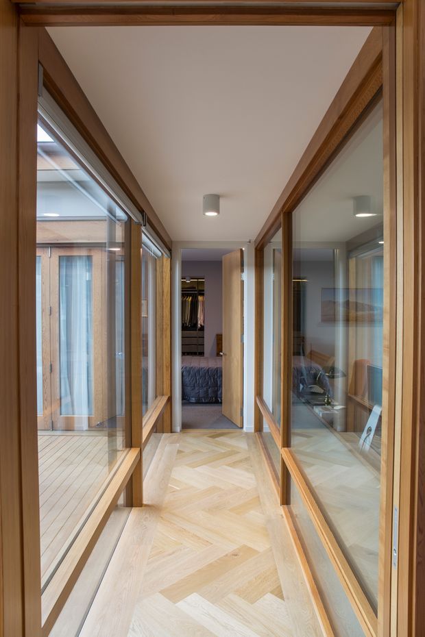 A narrow, bright interior corridor lined with extensive timber-framed glazing on both sides. The herringbone timber floor leads toward an open bedroom, while the glass walls reflect a compact sitting or workspace area. A timber-clad courtyard or glazed outdoor space is visible to the left. The corridor has a simple white ceiling with recessed cylindrical lighting and warm natural wood detailing throughout.