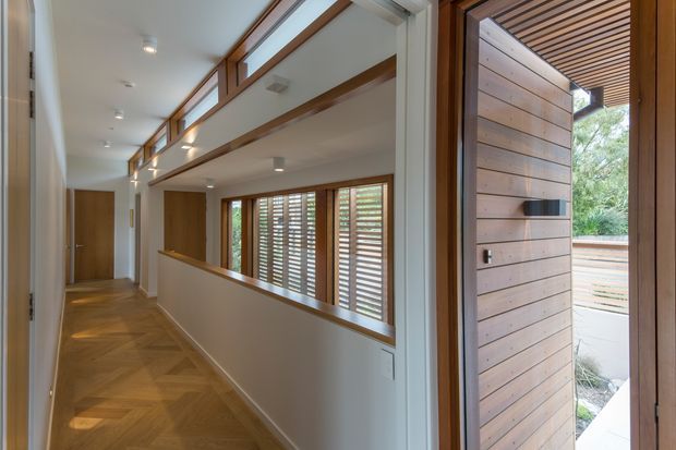 A bright, refined residential hallway viewed toward an open timber entry door. The corridor features warm herringbone timber flooring, white walls and ceiling, recessed downlights, timber-framed internal glazing with horizontal louvered shutters, and exposed timber beams above. Through the doorway, a covered exterior area with a slatted timber ceiling and horizontal timber-clad wall is visible.