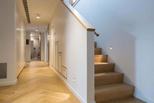 Bright contemporary residential hallway with a timber herringbone floor leading toward a bathroom or utility area, alongside an open timber staircase. The staircase has solid wood treads, a white side wall, and a simple timber handrail, with skylight-like glazing above. White walls, recessed lighting, built-in wall panels, and low-level step lights create a clean, refined interior.