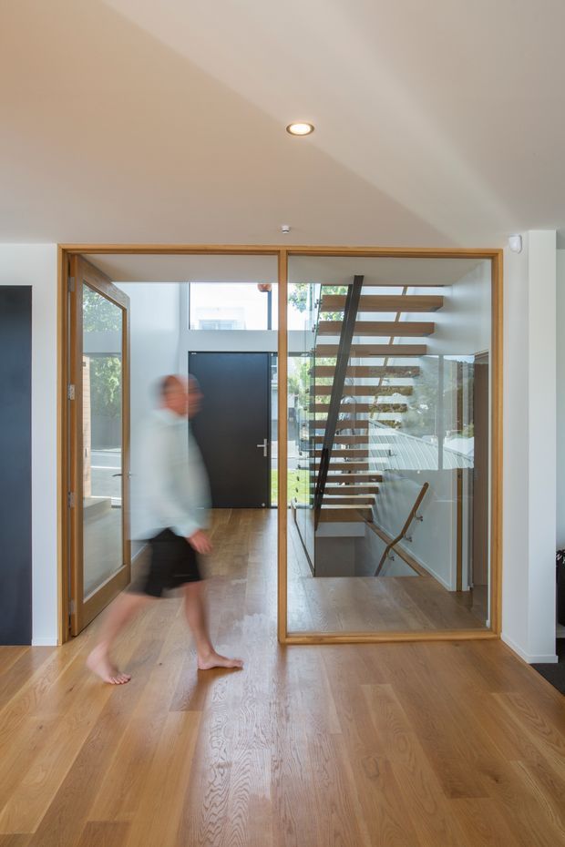 Bright contemporary interior hallway or landing with wide light timber flooring, a large timber-framed glazed partition, and views through to a black entry door and open-riser timber staircase. The space has white walls and ceiling, recessed lighting, abundant natural light, and a clean minimalist architectural composition. A blurred person is walking through the foreground.