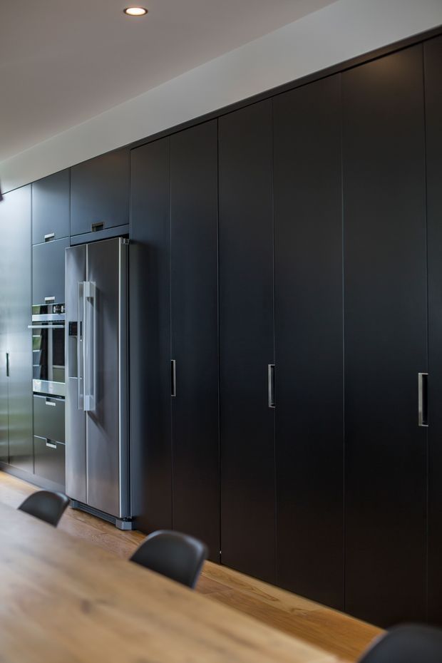 A contemporary kitchen with a full-height wall of sleek matte-black cabinetry, integrated dark storage, and a stainless-steel French-door refrigerator. A timber dining table and dark chairs occupy the foreground, with recessed ceiling lighting and warm timber flooring adding contrast.