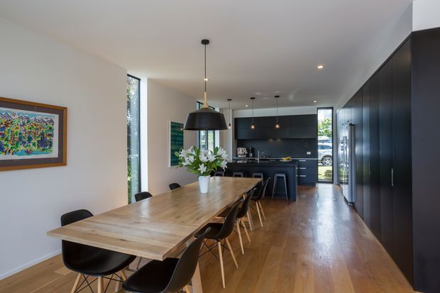 A contemporary open-plan dining and kitchen interior. A long light timber dining table with black chairs occupies the foreground, while a sleek dark kitchen with extensive floor-to-ceiling cabinetry, island seating, integrated appliances and pendant lighting runs along the right and rear. White walls, warm timber flooring, large windows and a framed colourful artwork create a bright, refined space.