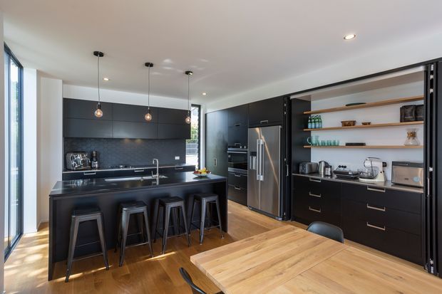 A contemporary open-plan kitchen and dining area with matte dark cabinetry, a central island with four bar stools, integrated stainless-steel appliances, a dark tiled splashback, open timber shelves, and a large timber dining table. Natural light enters through full-height glazing on the left, creating a balanced, refined interior.