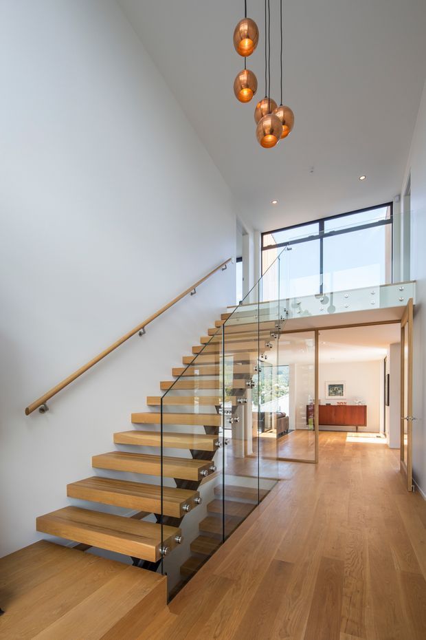 A bright contemporary interior hallway and stairway with a floating timber staircase, clear glass balustrades, and a timber handrail. The open stair rises beside a double-height void, with a timber floor extending through the corridor toward a living area. Large black-framed windows bring in abundant daylight, while a cluster of suspended amber pendant lights adds a warm focal point.