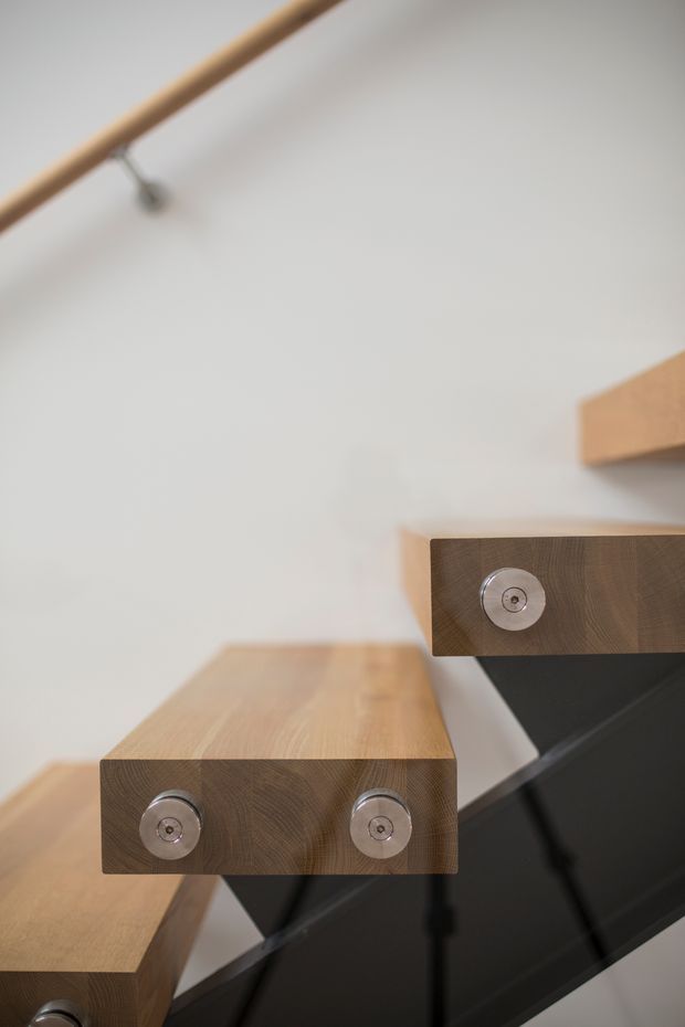 Close-up architectural detail of a modern floating staircase with thick solid timber treads, exposed circular metal fixings, and a dark black structural stringer or support. The stair rises diagonally through a minimal white interior, with a slim timber handrail visible above.