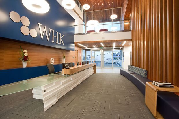 A contemporary commercial reception lobby with a prominent WHK feature wall, layered white reception desk, timber wall panelling, vertical timber slats, patterned carpet, lounge seating, and a double-height glazed entrance. Circular pendant lights and a timber-lined ceiling add visual interest, while a mezzanine level is visible above the entry.