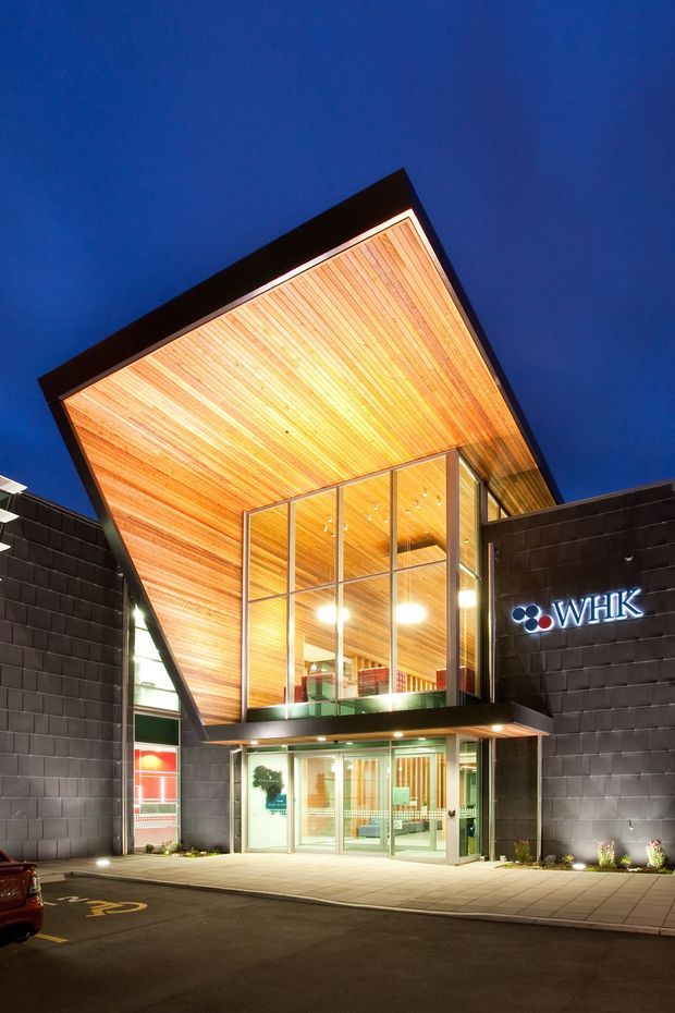 Night-time exterior photograph of a contemporary commercial building entrance. The facade is clad in dark rectangular masonry units, with a dramatic projecting canopy lined in warm timber, extending over a tall glazed lobby. Illuminated signage is mounted on the right facade, and a paved pedestrian entry fronts the building.